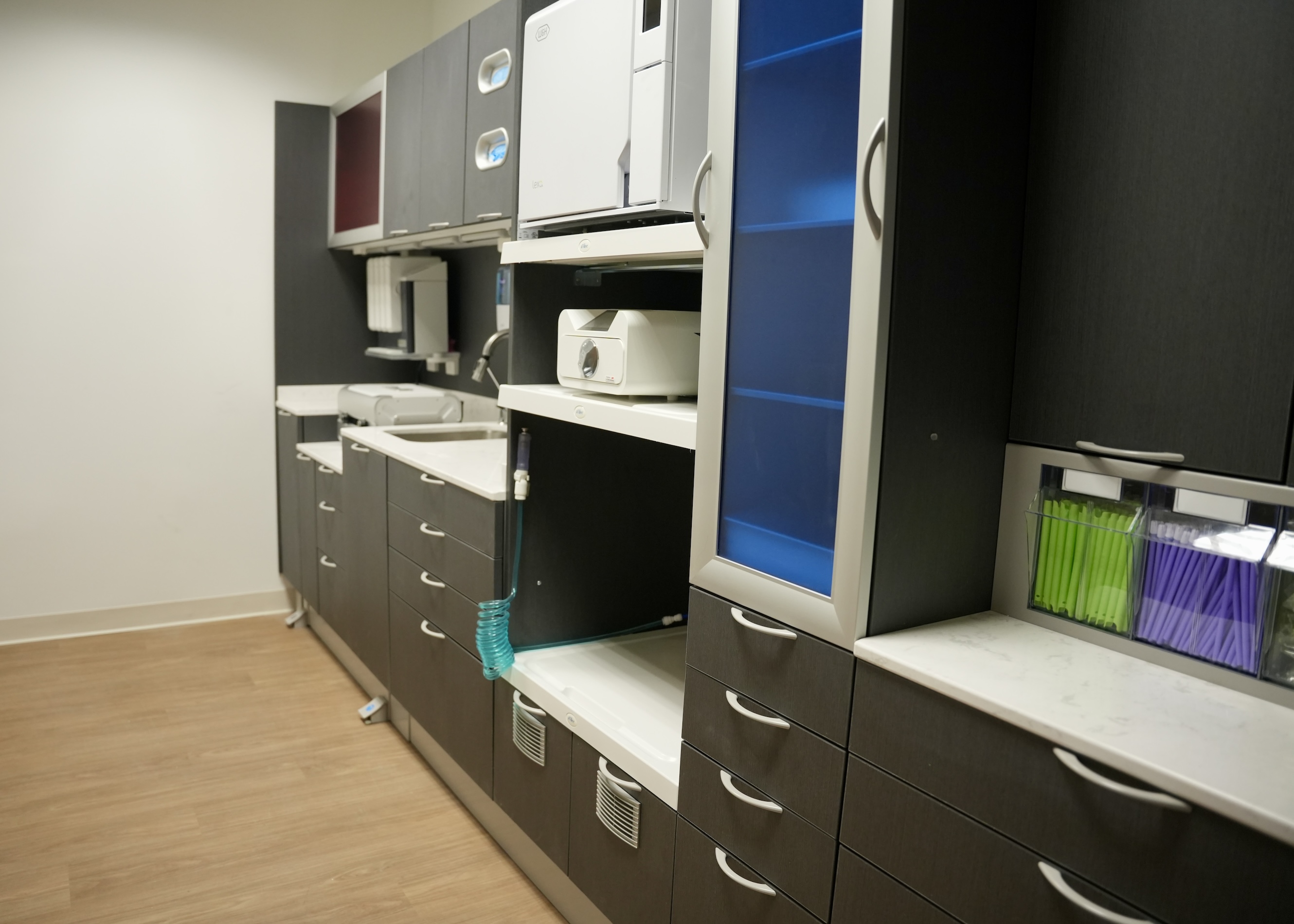 The image shows an office space with grey cabinets, white countertops, and various office equipment such as a printer, scanner, and computer monitors.