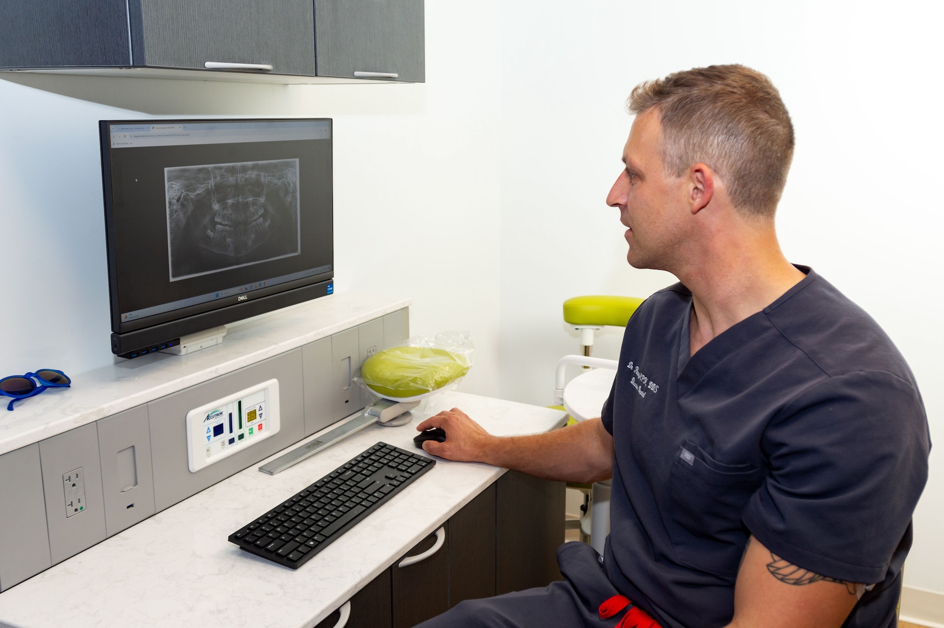 A man in scrubs sitting at a desk with a computer monitor displaying an X-ray image.