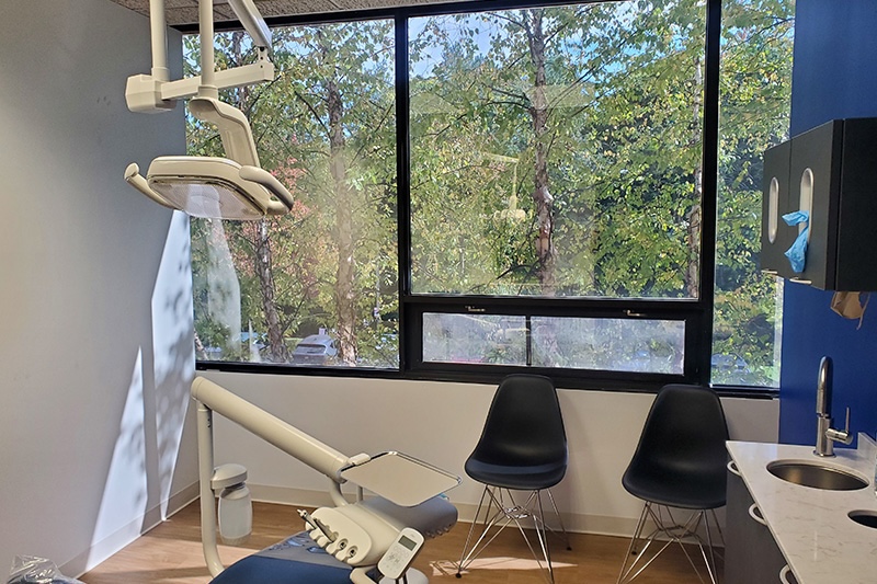 The image shows a dental office interior with a modern chair setup, a window with natural light, and various pieces of equipment typically found in such a setting.