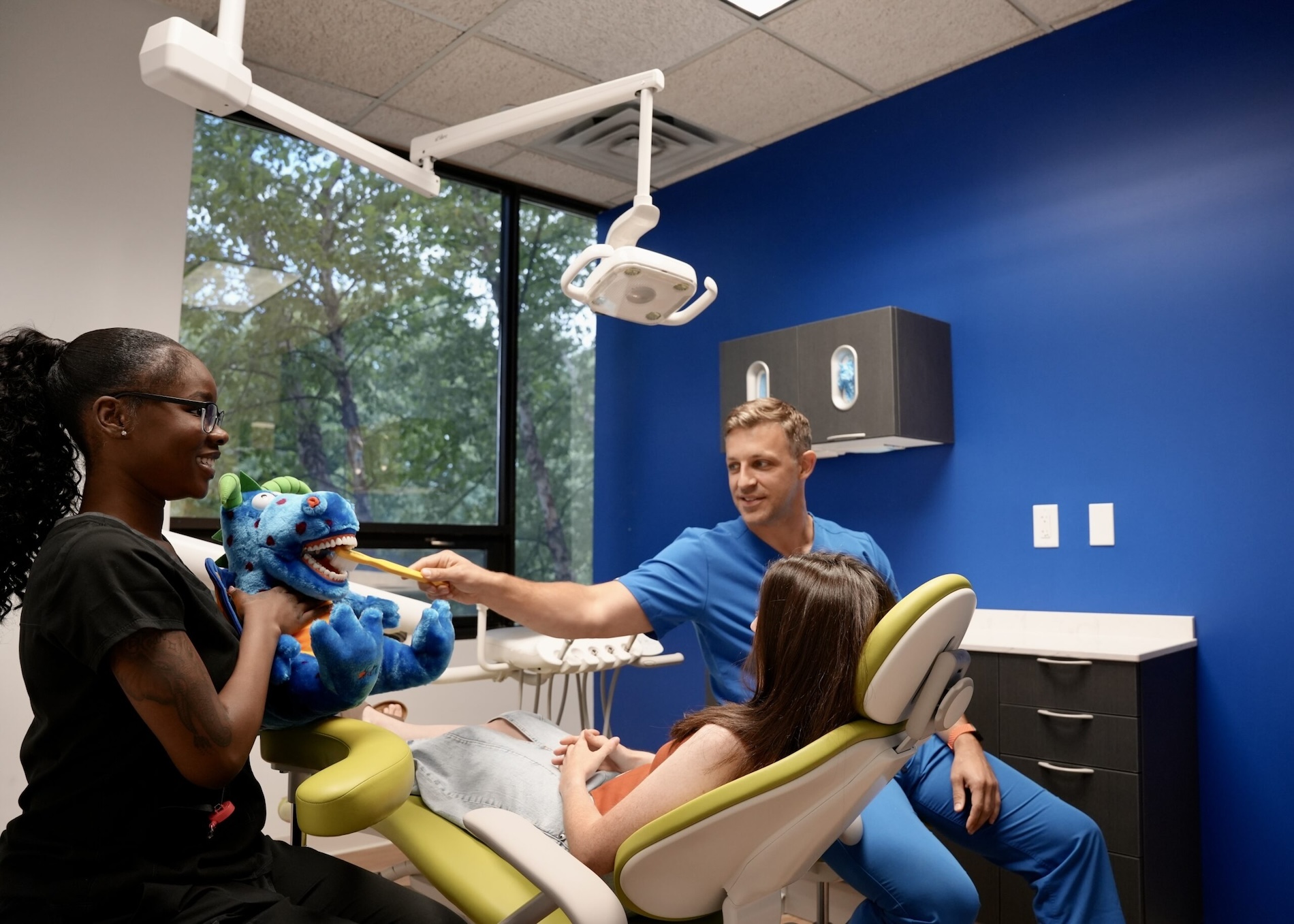 A man interacting with a child seated in a dental chair, with a stuffed animal and a woman standing behind them, all within a dental office setting.