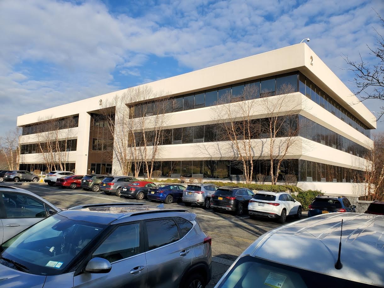 The image shows a modern multi-story building with a white exterior, large windows, and a flat roof, situated on a street corner with parked cars and a clear blue sky overhead.