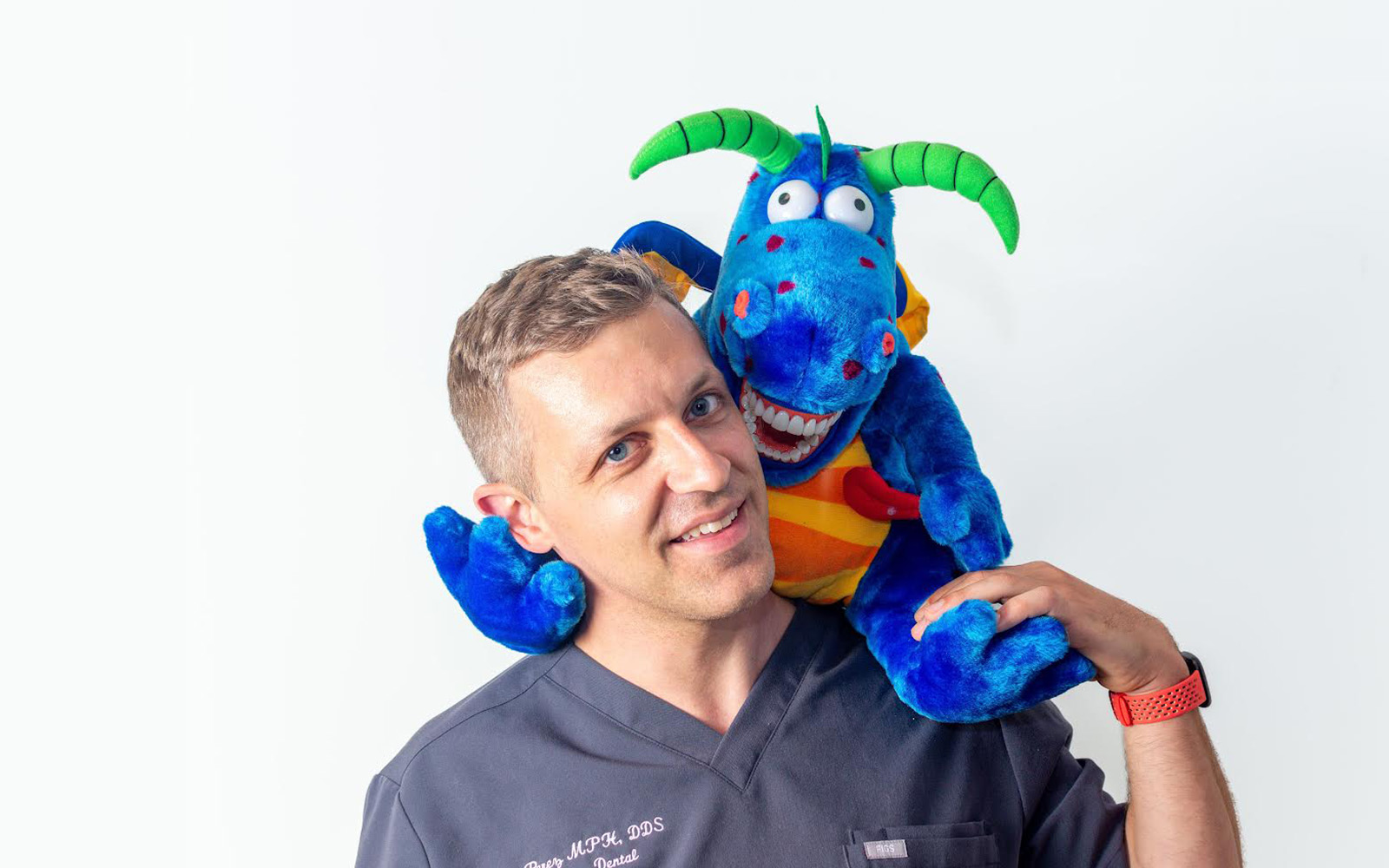 The image shows a man holding a blue stuffed animal with yellow eyes and an orange nose, who appears to be speaking or presenting, with a playful expression on his face.