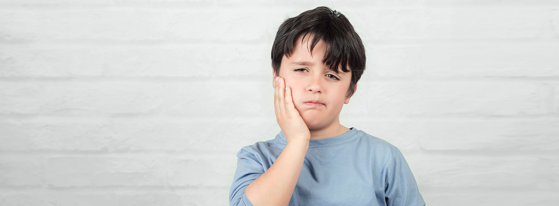 The image shows a young boy with his hand on his face, standing against a plain background.