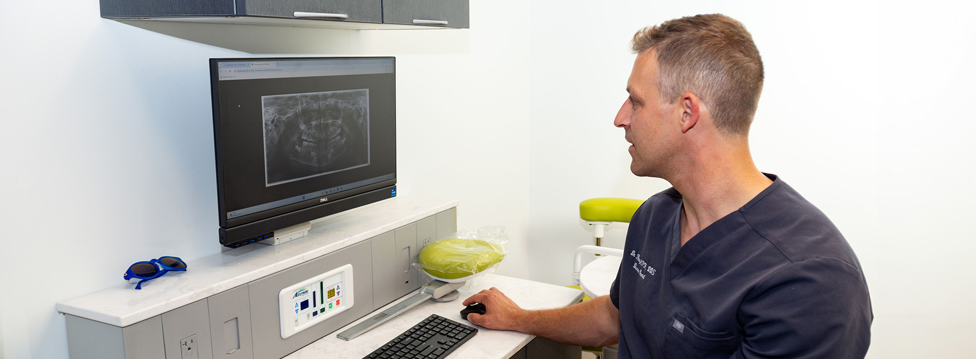 The image shows a man sitting at a desk with a computer monitor, keyboard, and mouse, in front of him is an X-ray machine.