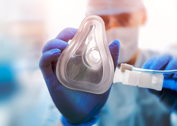 The image shows a person wearing gloves and a mask, holding a transparent medical device with a clear plastic casing, which appears to be a ventilator or respiratory aid, against a blurred background that suggests a laboratory setting.