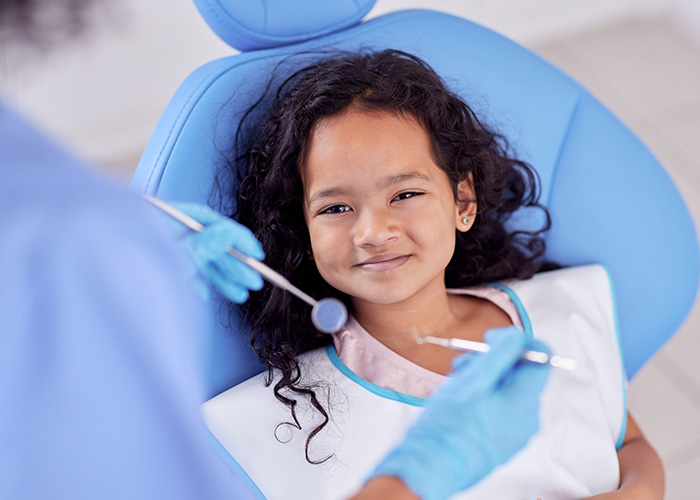 A young girl sitting in a dental chair with a smile on her face, receiving dental care from a professional wearing protective gloves and a mask, with a warm and caring atmosphere.