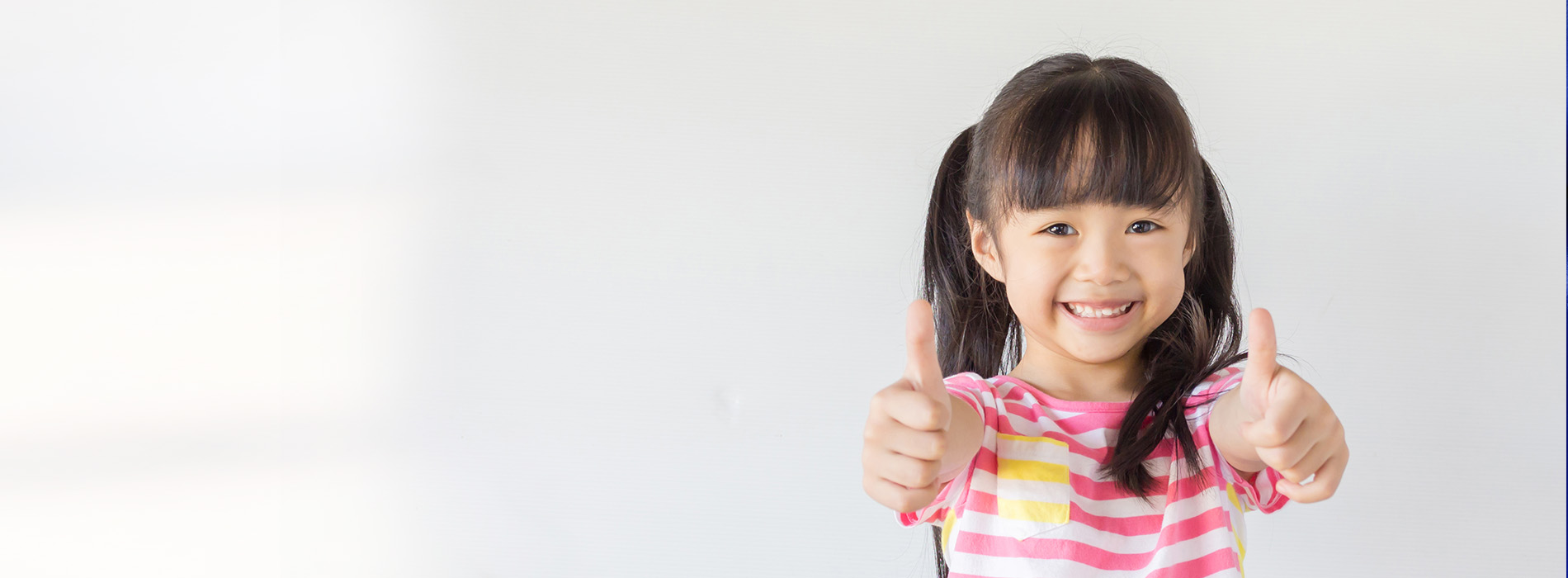 A young girl with dark hair smiling at the camera, giving a thumbs-up gesture.