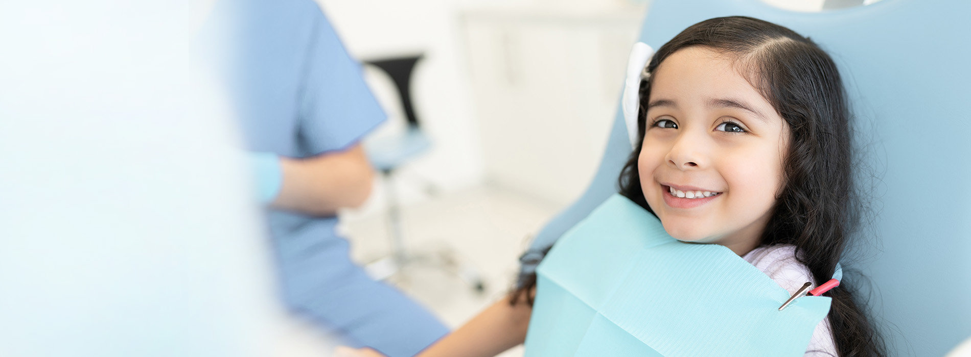 A young girl sitting in a dental chair with a smiling expression, wearing a protective blue bib, while a dental professional is behind her.