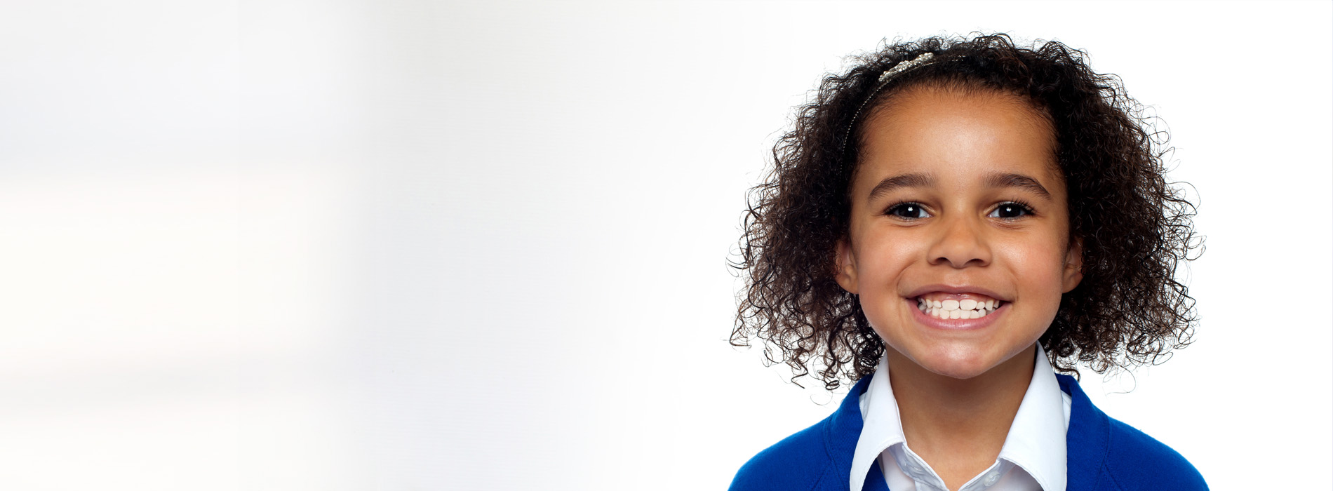 A young child with curly hair smiling at the camera, standing against a plain background.