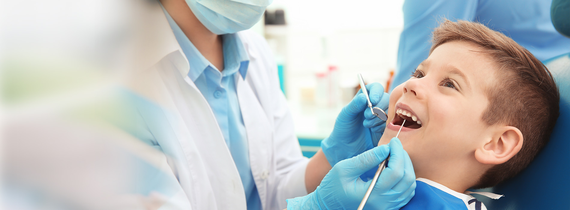 The image shows a dental professional in a white lab coat attending to a young child with a big smile, both wearing blue face masks and sitting in a dental chair, with a blurred background suggesting a medical setting.