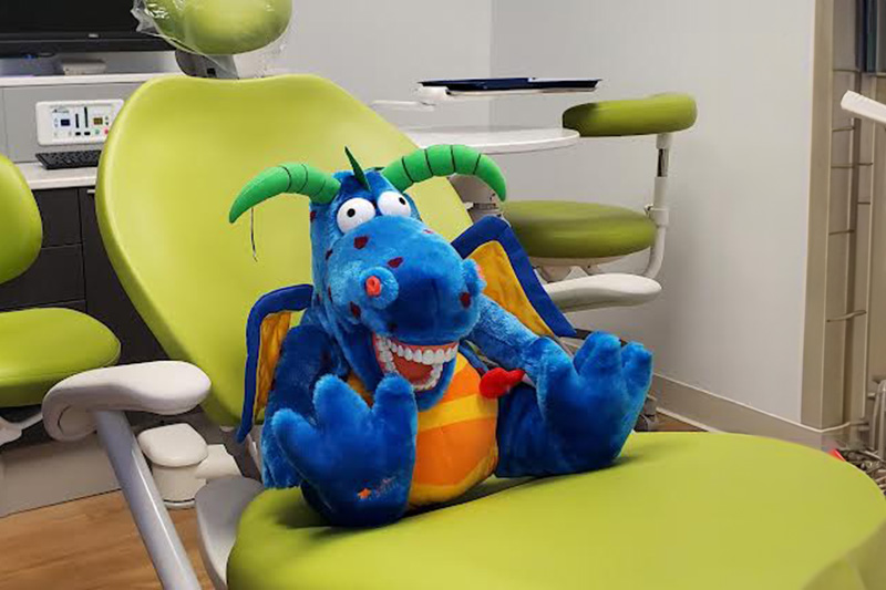 A plush blue dragon with a green horn sits on a dental chair in a dental office setting.